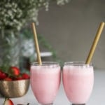 two glasses of strawberry lassi drink side by side with bamboo straw dipped inside alongside a bowl of fresh strawberries with white flowers in the background