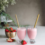 two glasses of strawberry lassi drink side by side with bamboo straw dipped inside alongside a bowl of fresh strawberries with white flowers in the background