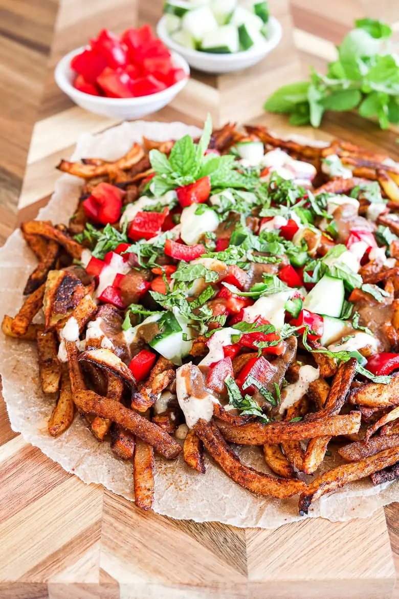 Spicy oven baked french fries loaded with peppers, cucumber and dressings with chopped herbs on a wooden board