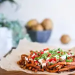 Spicy oven baked french fries loaded with peppers, cucumber and dressings with chopped herbs on a wooden board with raw potatoes and plants in the background