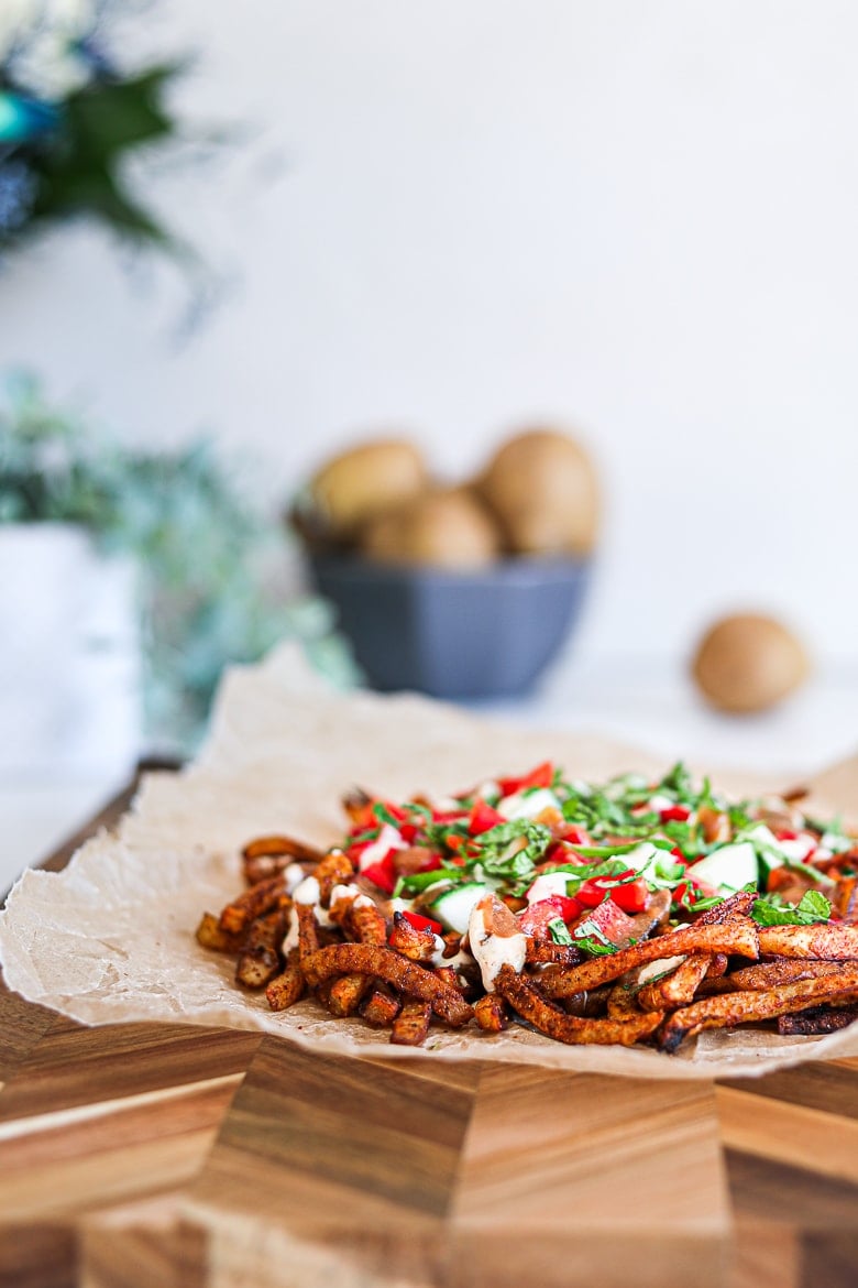 Spicy oven baked french fries loaded with peppers, cucumber and dressings with chopped herbs on a wooden board with raw potatoes and plants in the background