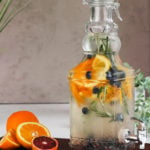 Perspective image of a pitcher containing a water infusion made with orange slices, blueberries, sprigs of rosemary displayed on a wooden board with oranges and blueberries on the side for decoration