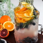 close up angled shot of a pitcher containing a fruit water infusion with orange slices, blueberries and sprigs of rosemary placed on a wooden board