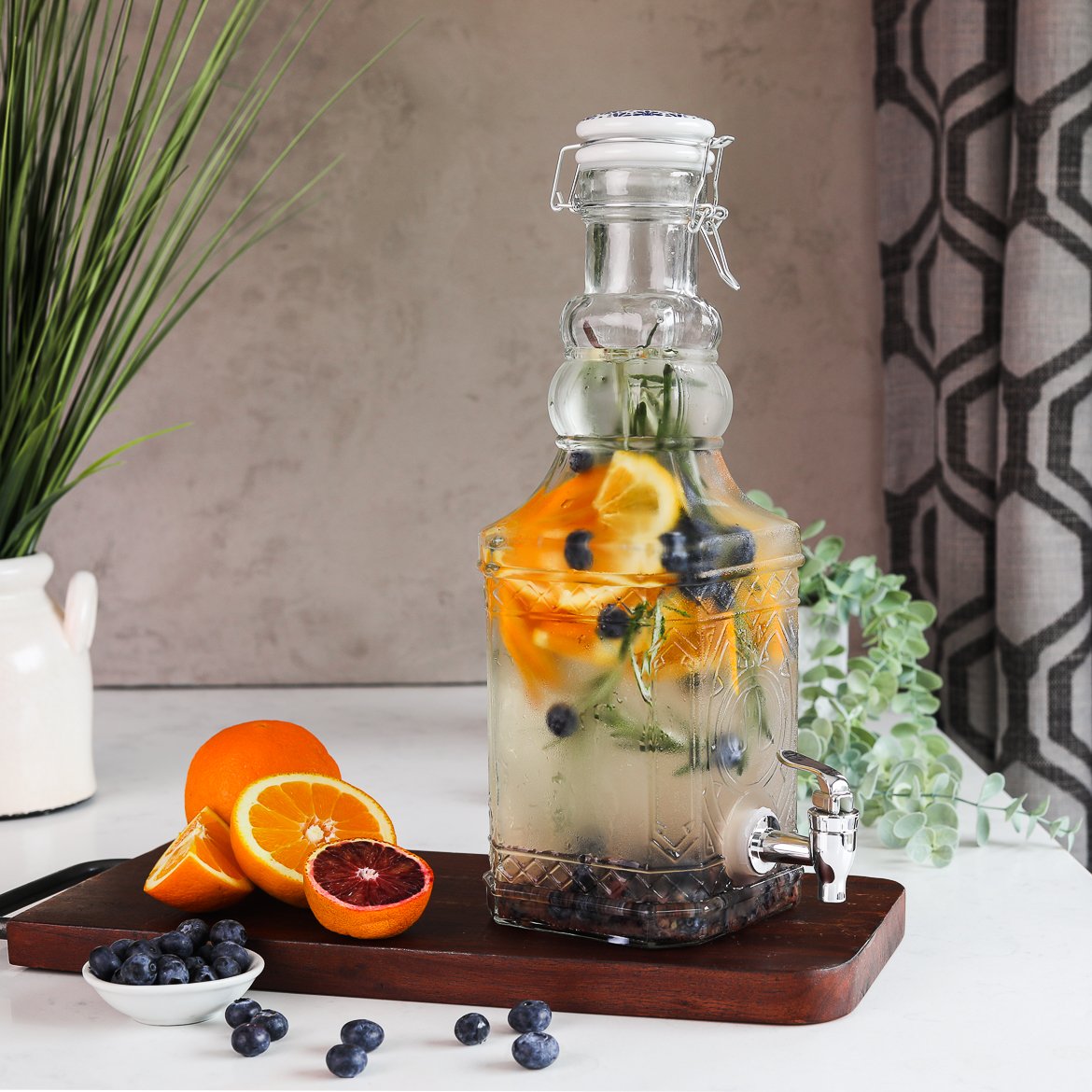 A pitcher containing a blueberry orange water infusion with springs of rosemary, displayed on a wooden board with fresh oranges and blueberries next to it