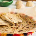 a colourful traditional South Asian straw basket containing a pile of parathas (Indian flat bread) with one paratha ripped in half. A rolling pin, a green bowl filled with chapati flour and balls of dough in the background