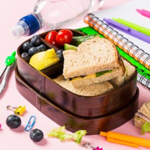 Wooden lunch box with sandwiches, vegetables and fruits on pink background and school stationery. Childrens eating concept.