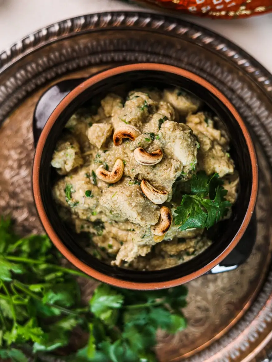 a clay pot with creamy chicken korma garnished with cilantro on a round rustic tray with a bunch of cilantro on the side