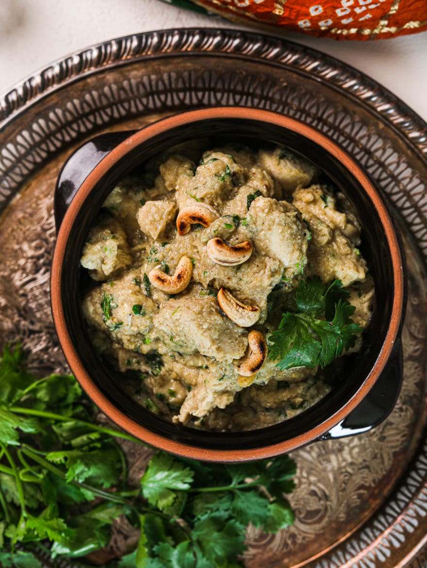 a clay pot with creamy chicken korma garnished with cilantro on a round rustic tray with a bunch of cilantro on the side