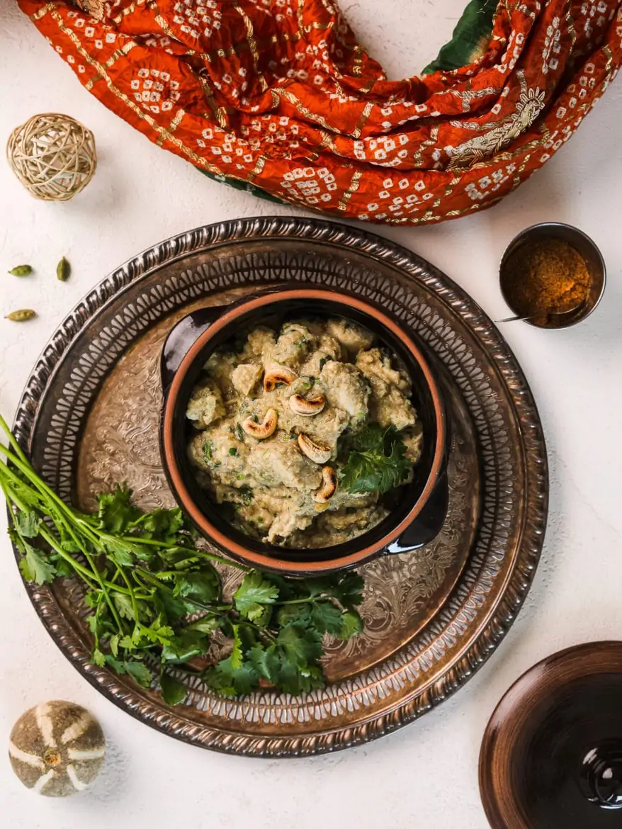 a clay pot with creamy chicken korma garnished with cilantro on a round rustic tray with a bunch of cilantro on the side and a traditional Indian scarf on the side - flatlay