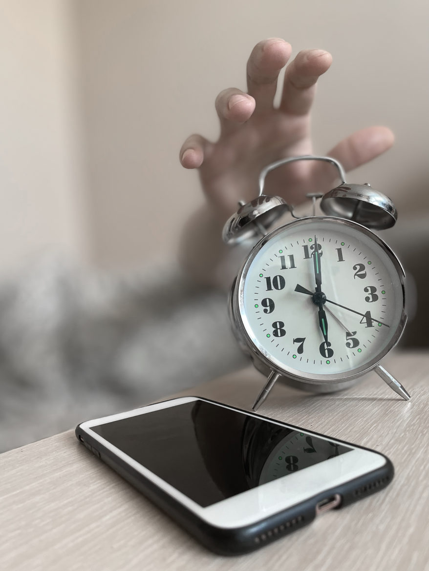 a hand reaching out for an alarm clock on a table with a cell phone next to it.