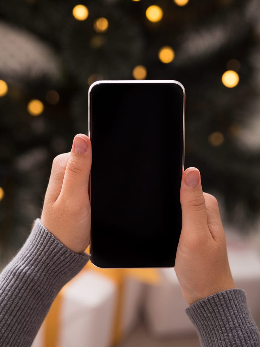 Searching for gifts. Cellphone in woman hands in over background with Christmas tree and presents, blank screen as natural ways to fall asleep