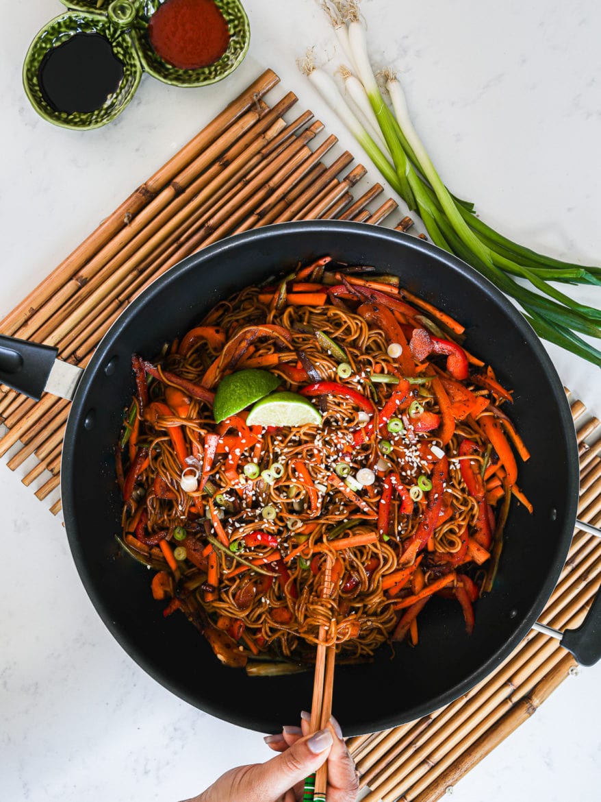 a wok with veg noodles topped with lime slices and sesame seeds - with someone holding a portion of noodles with chopsticks. Wok placed on a bamboo base with spring onions on the side