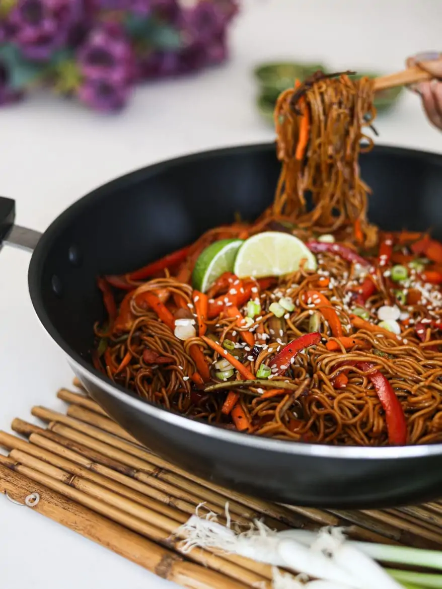 a wok with veg noodles topped with lime slices and sesame seeds - with someone holding a portion of noodles with chopsticks in the background - perspective shot