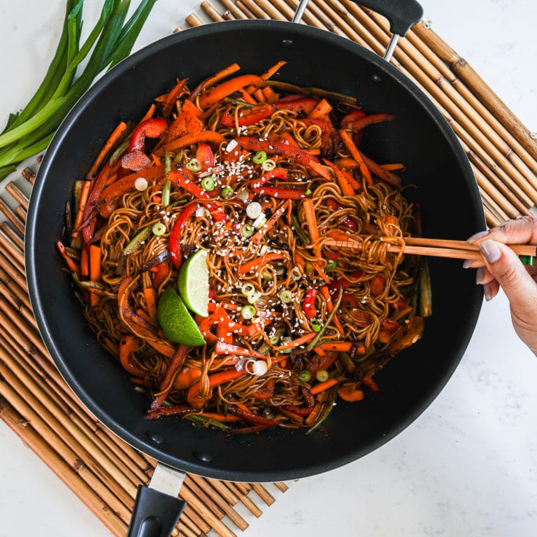 a wok with veg noodles topped with lime slices and sesame seeds - with someone holding a portion of noodles with chopsticks. Wok placed on a bamboo base with spring onions on the side