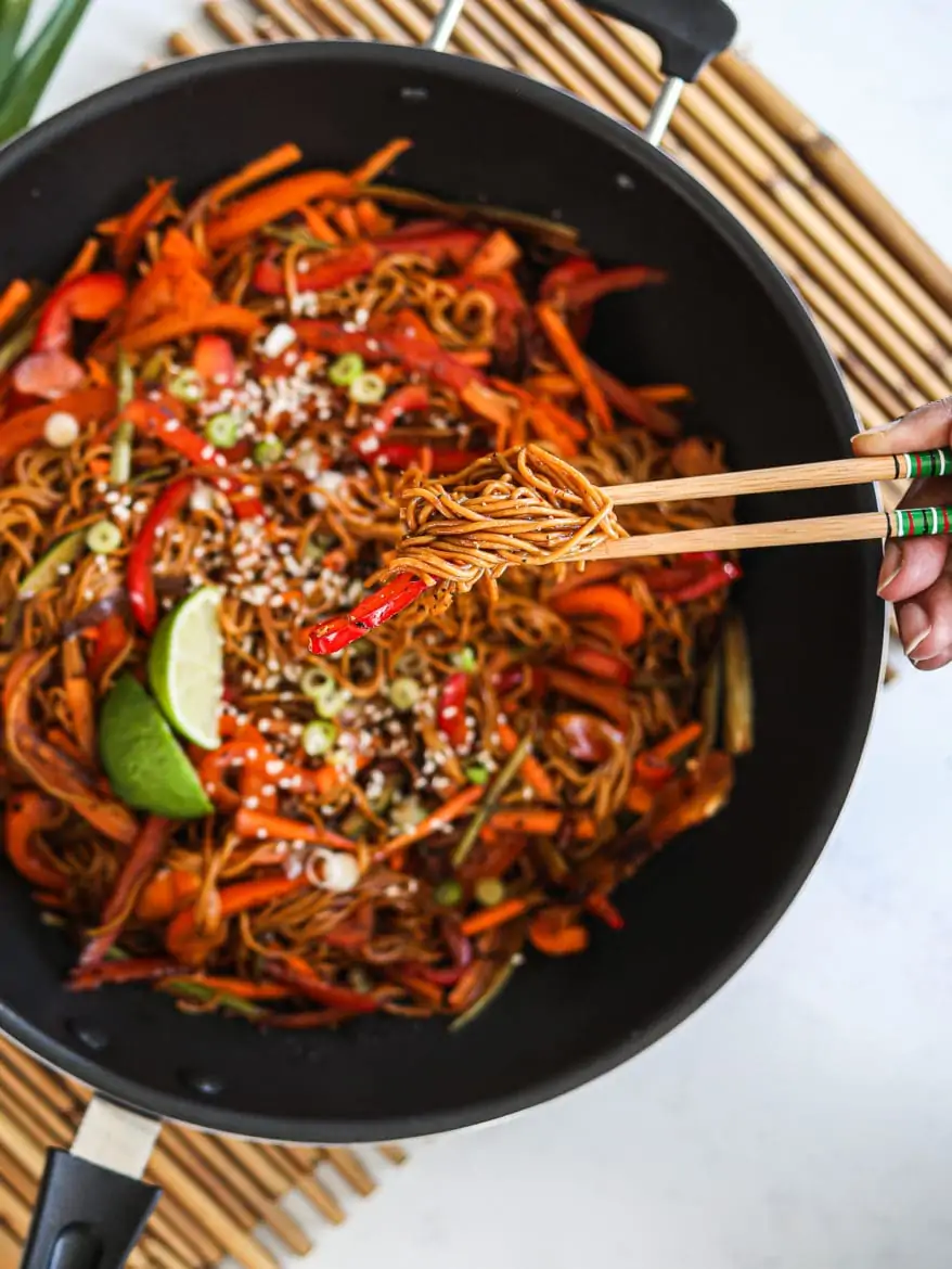 a wok with veg noodles topped with lime slices and sesame seeds - with someone holding a portion of noodles with chopsticks