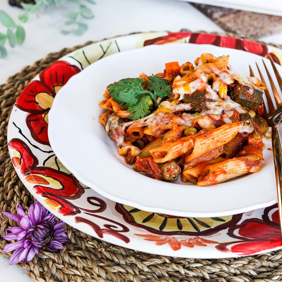 a plate with pasta bake made with penne pasta, okra and mixed vegetables. Garnished with cilantro. Styled on top of a floral plate on a straw mat.