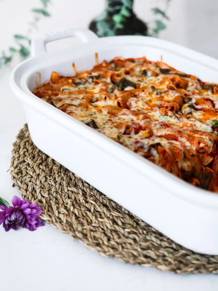 a white rectangular casserole dish with pasta bake topped with melted cheese on a brown straw mat with a green plant in the background