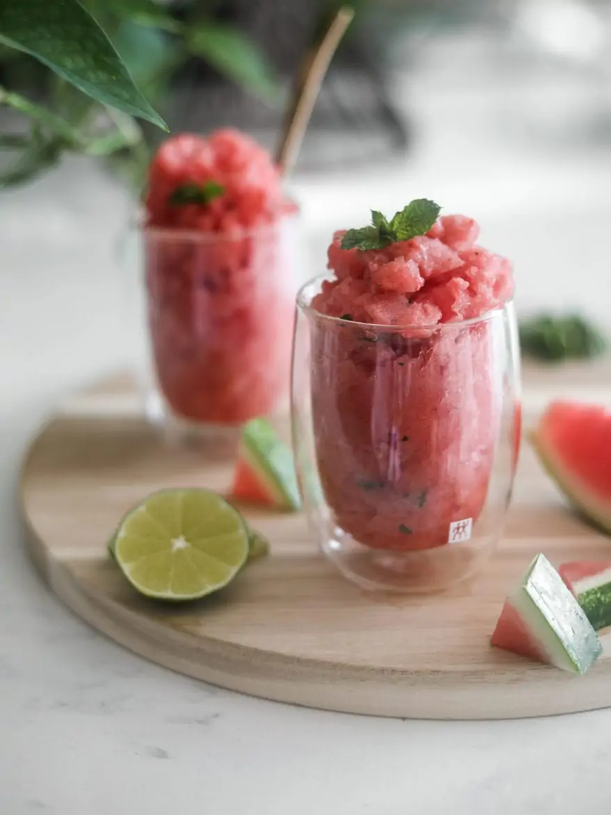 two glasses of watermelon slushie on a round wooden board with pieces of watermelon and lime slices close by with a green plant in the background