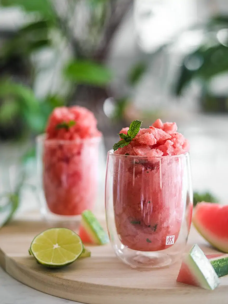 two glasses of watermelon slushie on a round wooden board with pieces of watermelon and lime slices close by with a green plant in the background