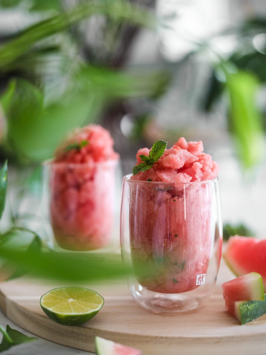 two glasses of watermelon slushie on a round wooden board with pieces of watermelon and lime slices close by and a green plant decoration