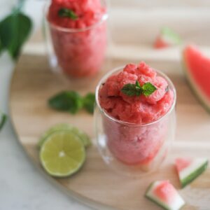 two glasses of watermelon slushie on a round wooden board with pieces of watermelon and lime slices close by
