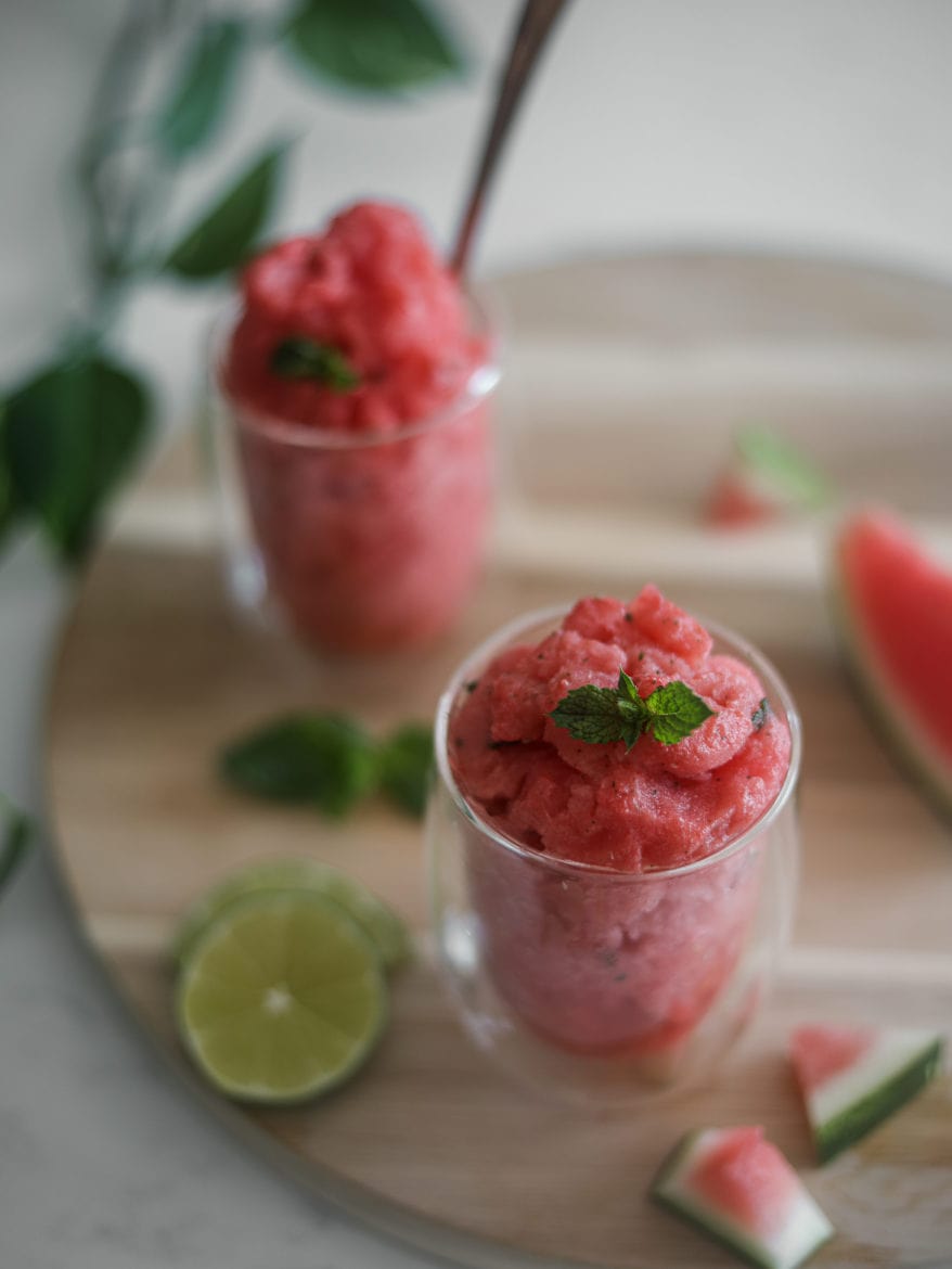 two glasses of watermelon slushie on a round wooden board with pieces of watermelon and lime slices close by