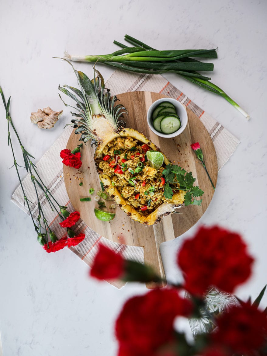 half a pineapple stuffed with Thai pineapple fried rice topped with a lime segment and cilantro on a round wooden board decorated with red flowers arranged around.