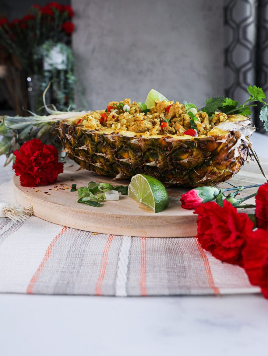 side shot of half a pineapple stuffed with Thai pineapple fried rice topped with cilantro on a round wooden board decorated with red flowers arranged around.