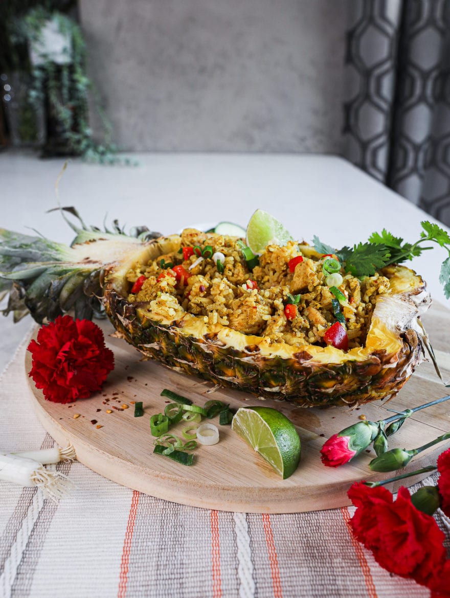 side shot of half a pineapple stuffed with Thai pineapple fried rice topped with a lime segment and cilantro on a round wooden board decorated with red flowers arranged around.