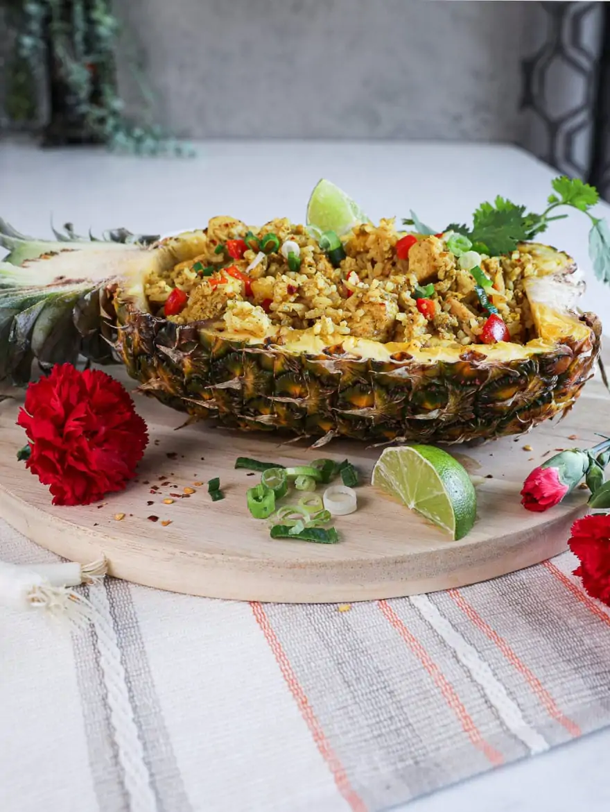 side shot of half a pineapple stuffed with Thai pineapple fried rice topped with a lime segment and cilantro on a round wooden board decorated with red flowers arranged around.