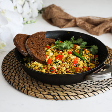 fry pan of vegan tofu scramble with red pepper cubes topped with cilantro and green chilli and two slices of pumpernickel bread placed on top