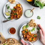 Top down down view of a vegetarian, vegan Indian Balti curry with cauliflower and pumpkin served with raita, naan bread and mango chutney and basmati rice on white background, female hands holding one plate