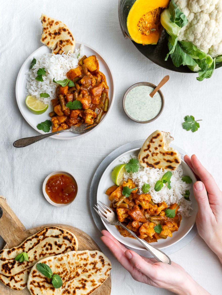 Top down down view of a vegetarian, vegan Indian Balti curry with cauliflower and pumpkin served with raita, naan bread and mango chutney and basmati rice on white background, female hands holding one plate