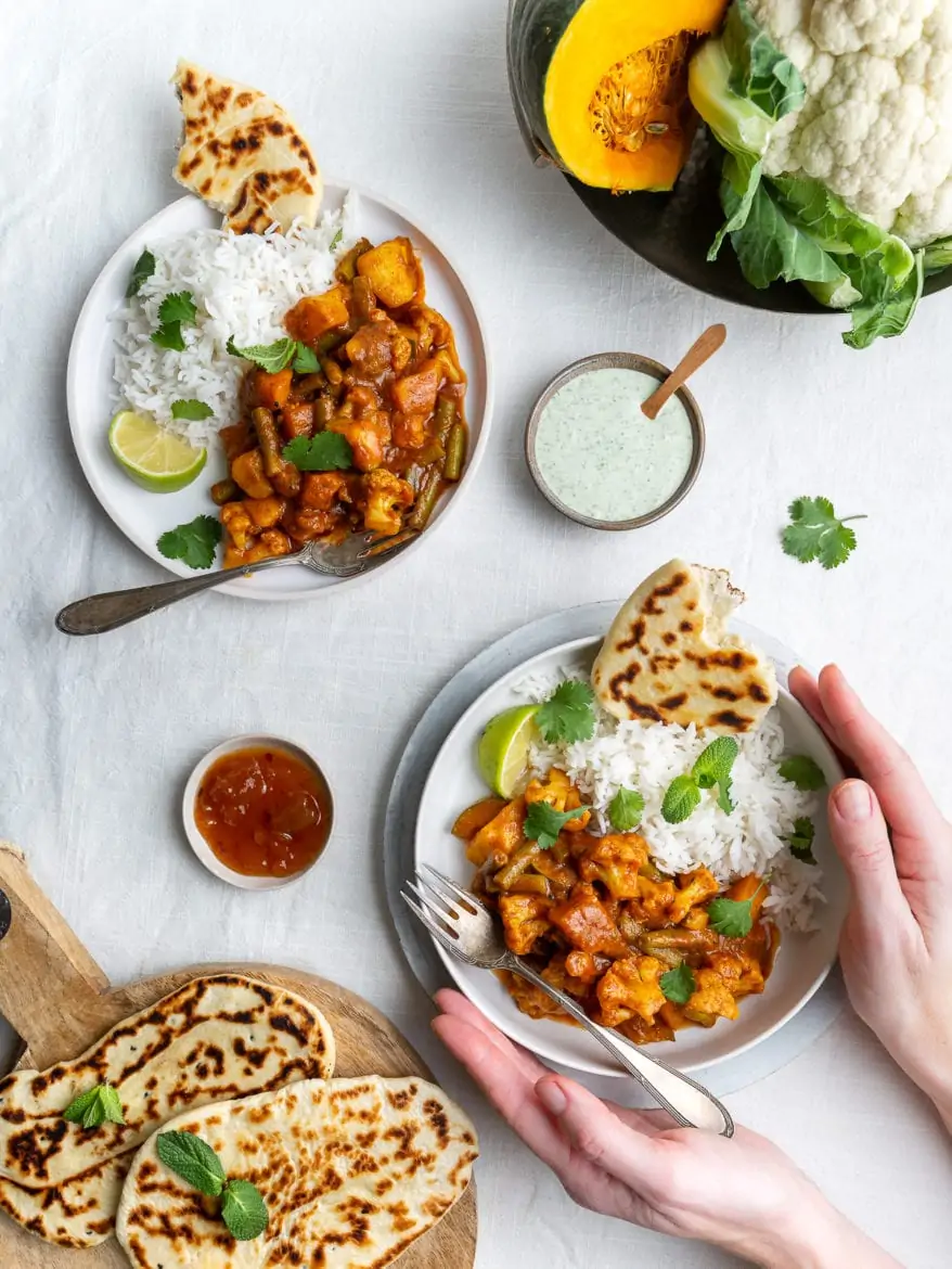 Top down down view of a vegetarian, vegan Indian Balti curry with cauliflower and pumpkin served with raita, naan bread and mango chutney and basmati rice on white background, female hands holding one plate