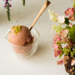 a small glass bowl of watermelon ice cream topped with crushed pistachios and a gold spoon standing in the bowl - flowers in the fore- and background.