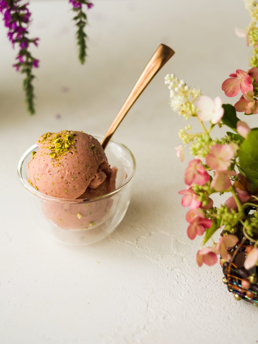 a small glass bowl of watermelon ice cream topped with crushed pistachios and a gold spoon standing in the bowl - flowers in the fore- and background.
