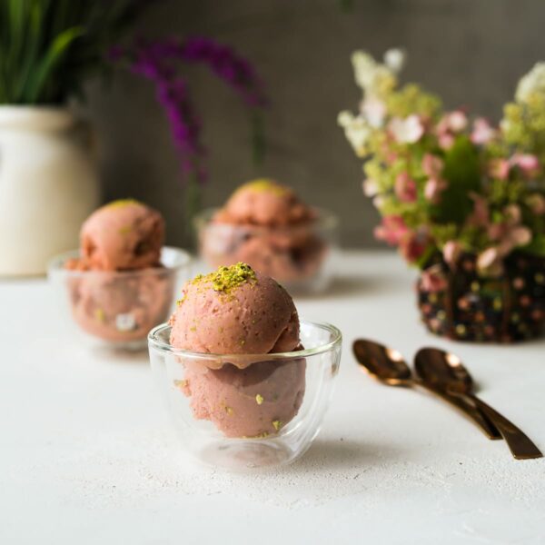 three bowls of vegan watermelon ice cream topped with pistachios with plants in the fore- and background. Two gold spoons nearby.