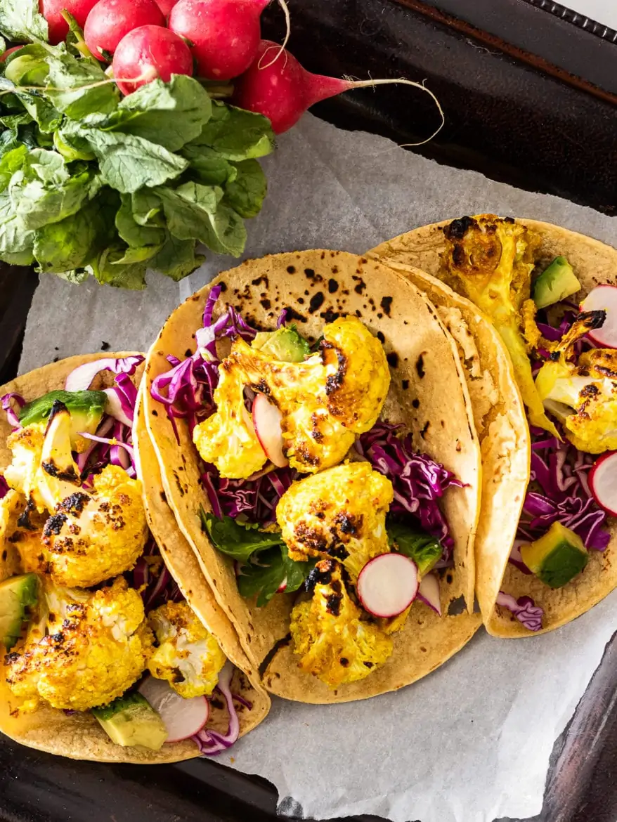 Tray with three corn cauliflower tacos with purple cabbage and avocado cubes