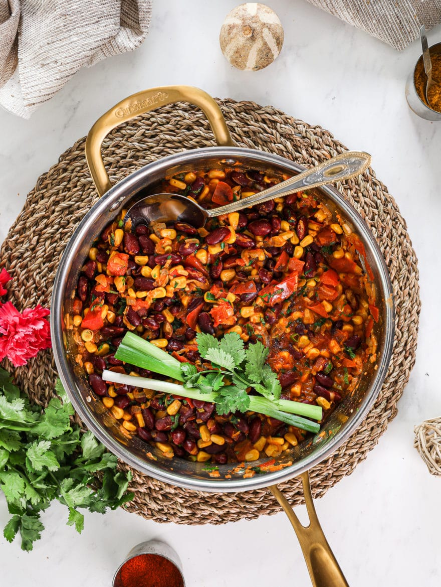 a pan pf githeri (kenyan beans and corn dish on a straw mat with herbs and flowers decorated around and a beige scarf styled close by. Close up flatlay shot.