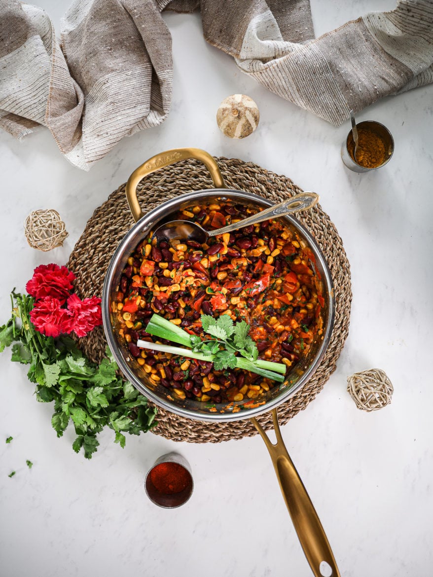 a pan pf githeri (kenyan beans and corn dish on a straw mat with herbs and flowers decorated around and a beige scarf styled close by. Flatlay.