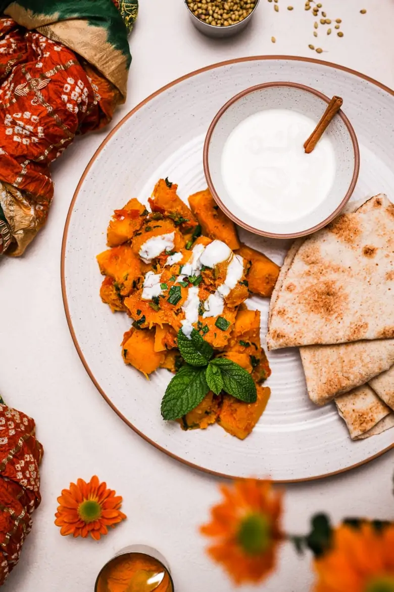Kadoo (Afghan Pumpkin Curry) presented on a white plate alongside a small bowl of yoghurt and pita bread on a white background with flowers and spices pots close by