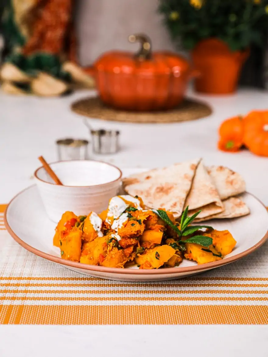 Kadoo (Afghan Pumpkin Curry) presented on a white plate alongside a small bowl of yoghurt and pita bread on a white background with two small aluminium bowls and a pumpkin shaped cocotte with a few orange flowers in the foreground