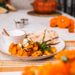 Kadoo (Afghan Pumpkin Curry) presented on a white plate alongside a small bowl of yoghurt and pita bread on a white background with two small aluminium bowls and a pumpkin shaped cocotte with a few orange flowers in the foreground