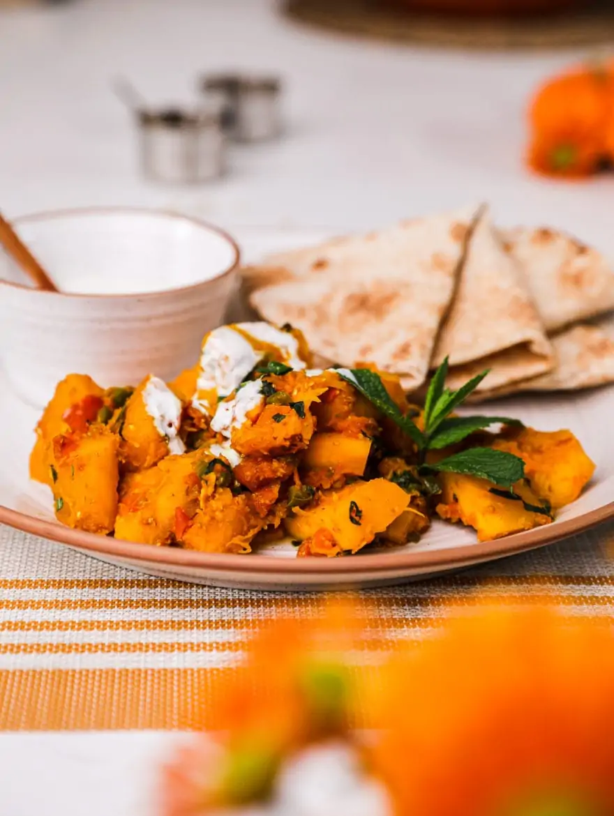 Kadoo (Afghan Pumpkin Curry) presented on a white plate alongside a small bowl of yoghurt and pita bread on a white background with two small aluminium bowls and few orange flowers in the foreground