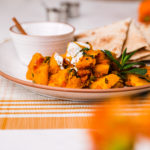 Kadoo (Afghan Pumpkin Curry) presented on a white plate alongside a small bowl of yoghurt and pita bread on a white background with two small aluminium bowls and few orange flowers in the foreground