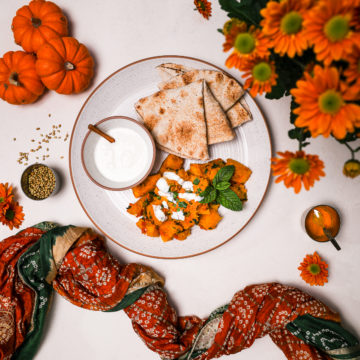 Kadoo (Afghan Pumpkin Curry) presented on a white plate alongside a small bowl of yoghurt and pita bread on a white background with a traditional scarf, small pumpkins and orange flowers all around.
