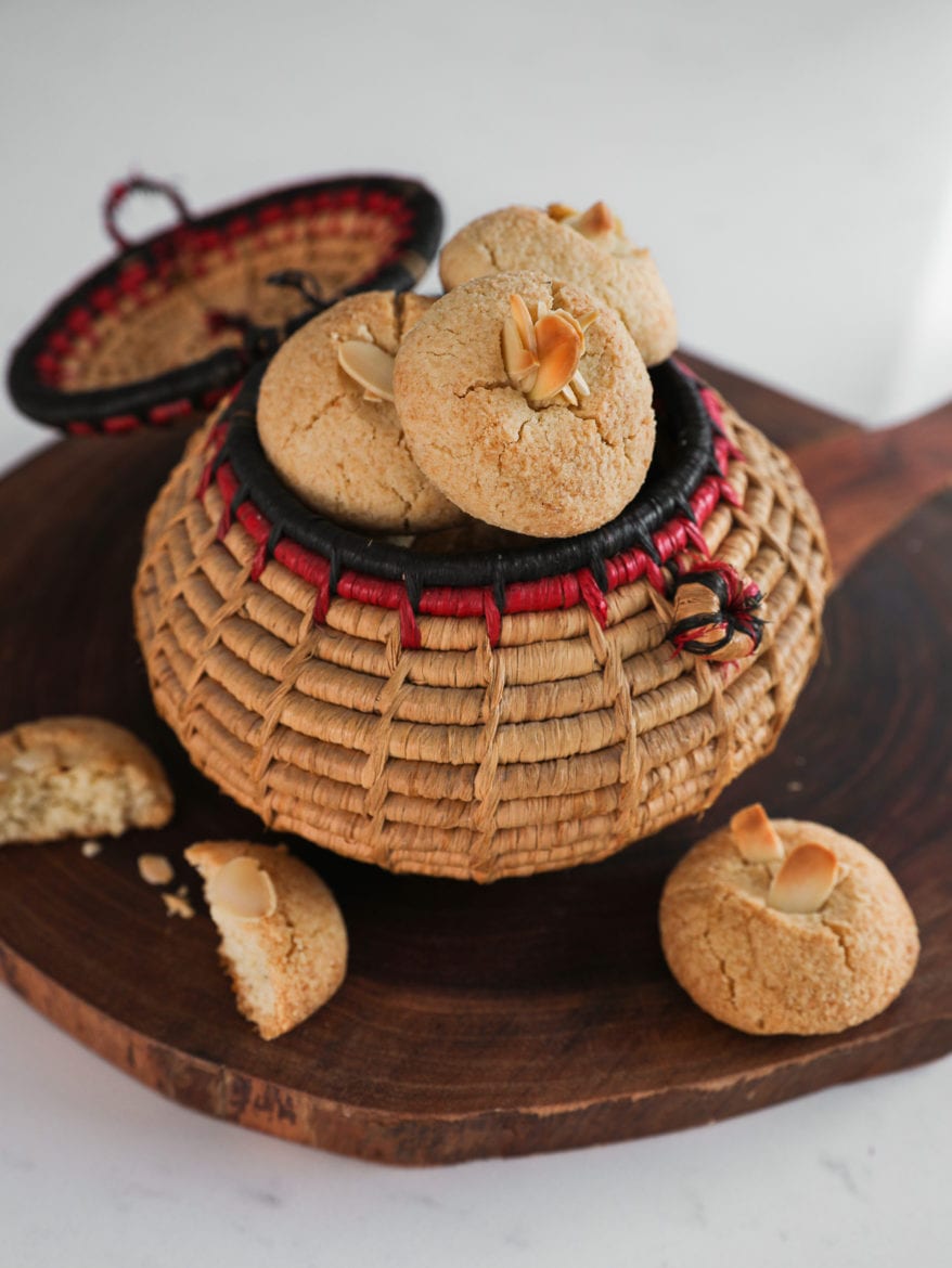 a traditional round basket filled to the brim with almond flour cookies topped with sliced almonds on a round wooden board