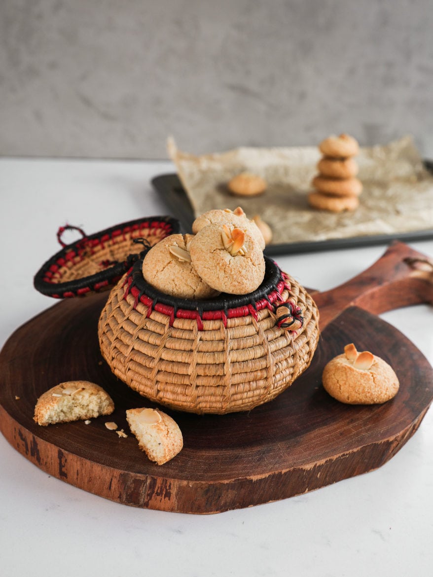 a traditional round basket filled to the brim with almond flour cookies topped with sliced almonds on a round wooden board with a baking sheet in the background with a pile of cookies on top