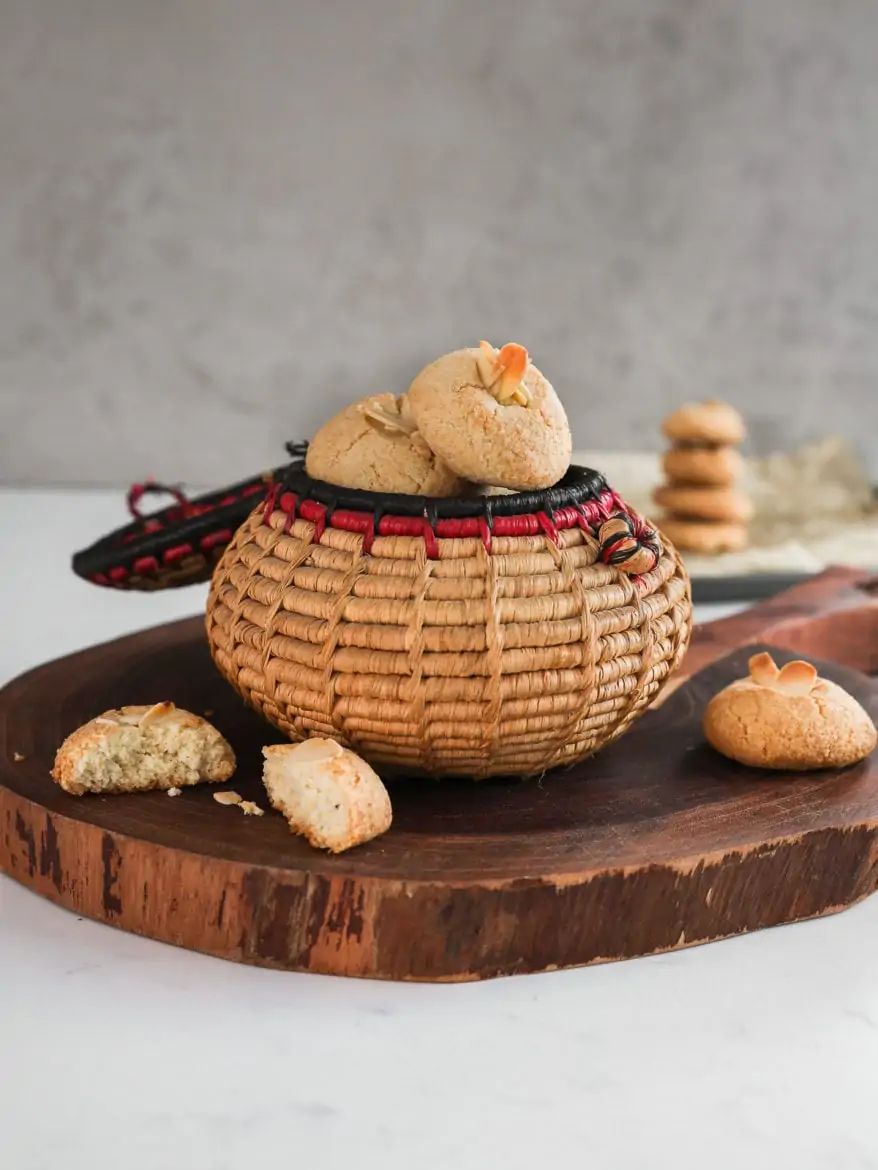 a close up shot of a traditional round basket filled to the brim with almond flour cookies topped with sliced almonds on a round wooden board with a baking sheet in the background with a pile of cookies on top