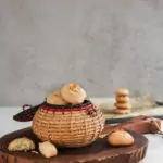 perspective shot of a traditional round basket filled to the brim with almond flour cookies topped with sliced almonds on a round wooden board with a broken cookie. There is also a baking sheet in the background with a pile of cookies on top